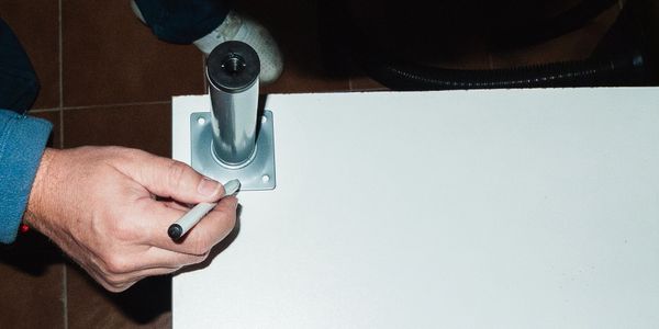A person marking a spot on a white surface near a metal table leg.