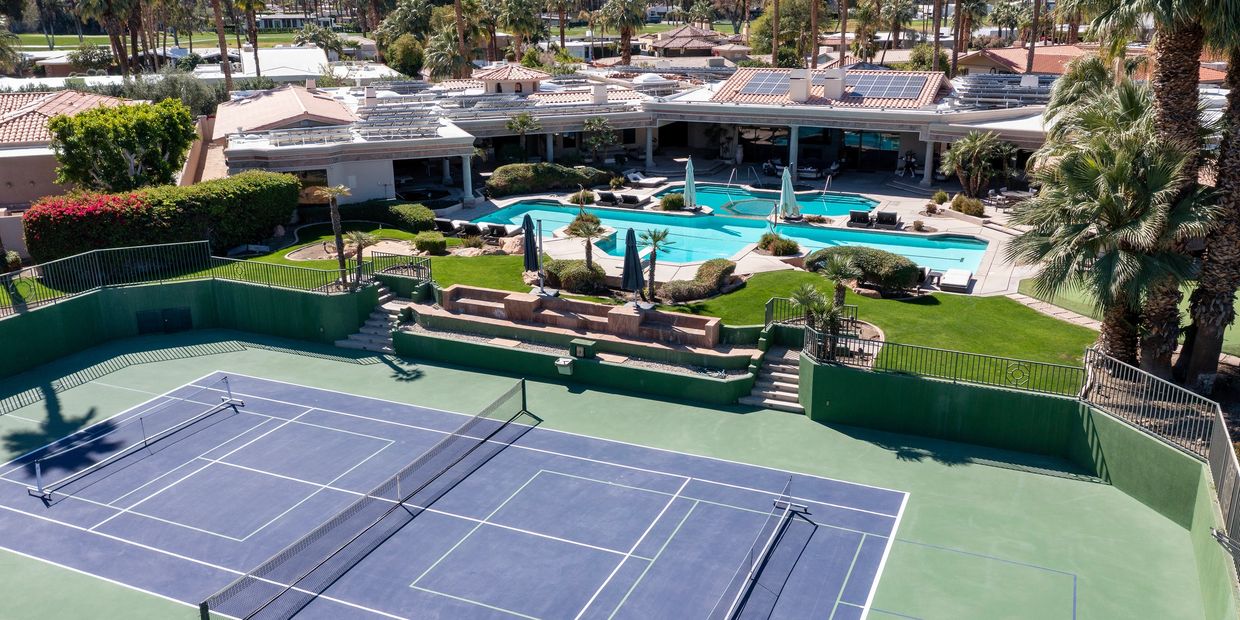 A tennis court next to a swimming pool and clubhouse in a sunny resort.