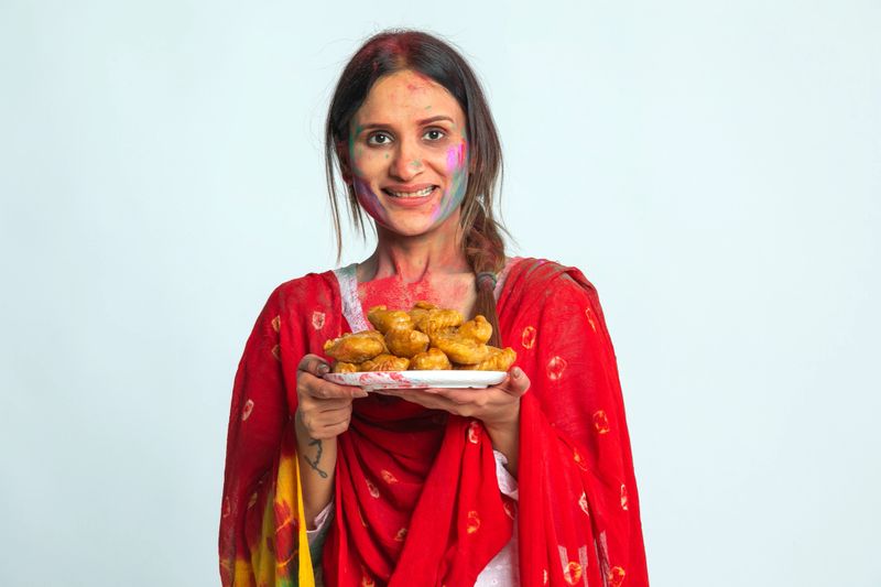 Portrait of excited beautiful woman holding delicious gujiyas while standing against white background during Holi festival