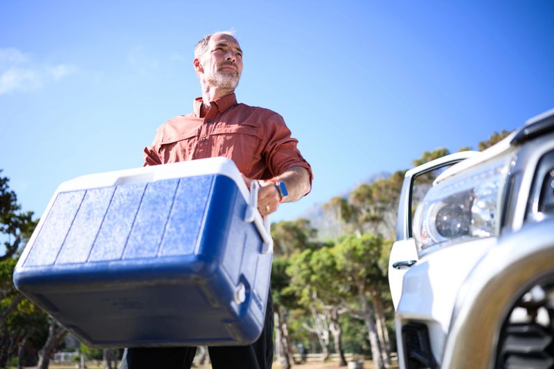Man unpacking camping crates and equipment from utility vehicle parked next to trees on a remote beach surrounded by mountains