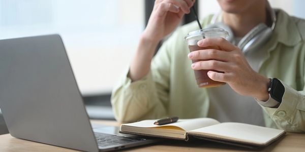 Person stirring an iced coffee while working on their laptop.  Also has a note pad out for reference