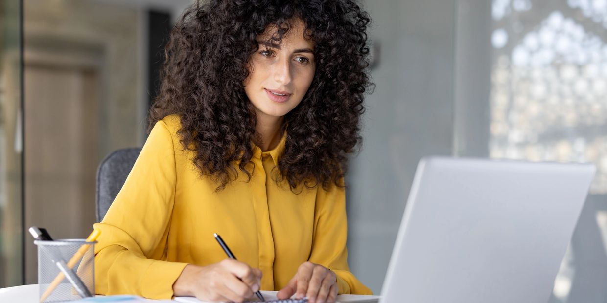 Woman in yellow shirt working at laptop and writing in notebook.