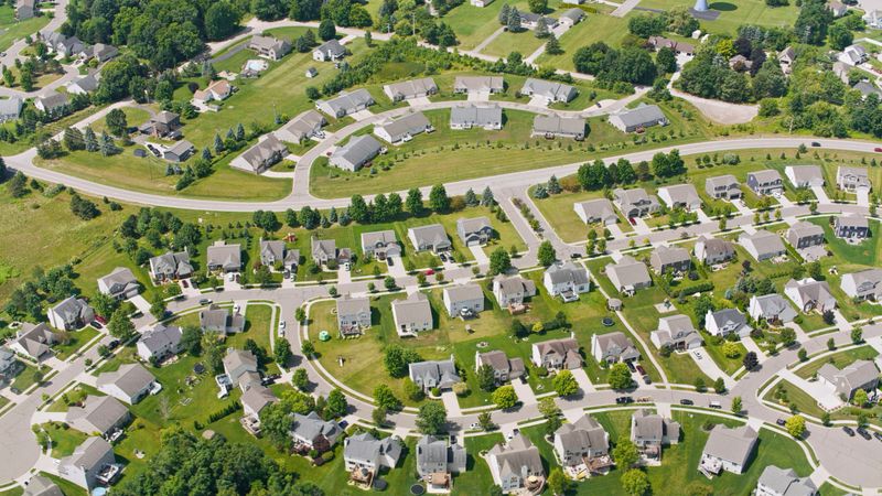 Aerial view of Lansing Michigan suburbs featuring neatly arranged homes, winding roads, and lush green lawns under a sunny sky. Peaceful residential setting.