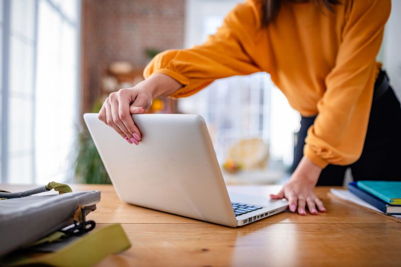 A young woman in a bright orange top takes a moment to unwind at home, closing her laptop in a stylish and comfortable living room. She radiates happiness and contentment.