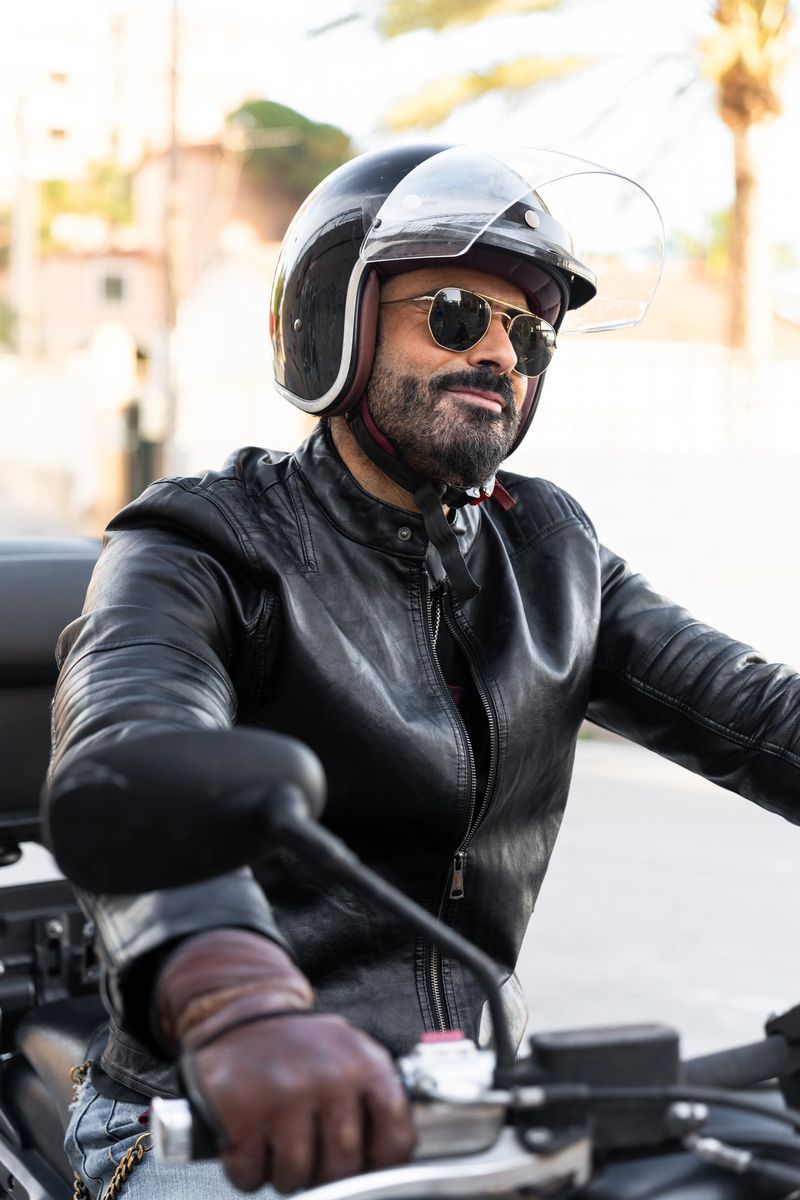 Confident mature male biker wearing sunglasses, helmet and leather jacket riding his motorcycle in the city