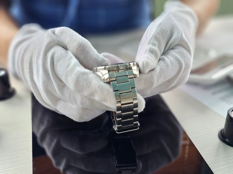A skilled watch technician examines a wristwatch under bright lights, wearing white gloves to handle the delicate piece carefully on a polished work surface.