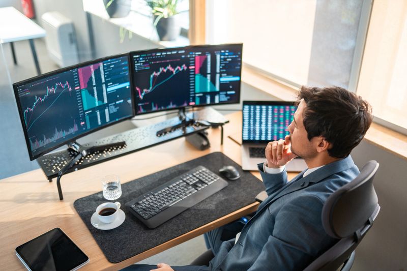A mid adult Asian stockbroker in a suit examines detailed financial charts on multiple monitors at a modern workspace, reflecting a focused and analytical demeanor.