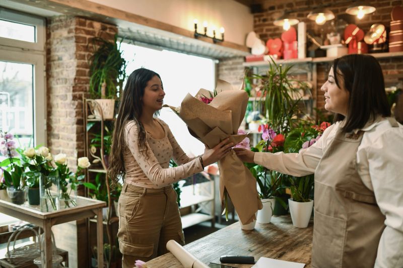 A woman florist hands a beautiful bouquet of flowers to a happy customer inside a cozy shop filled with vibrant plants and natural decor, capturing the atmosphere of gifting and personalized service.