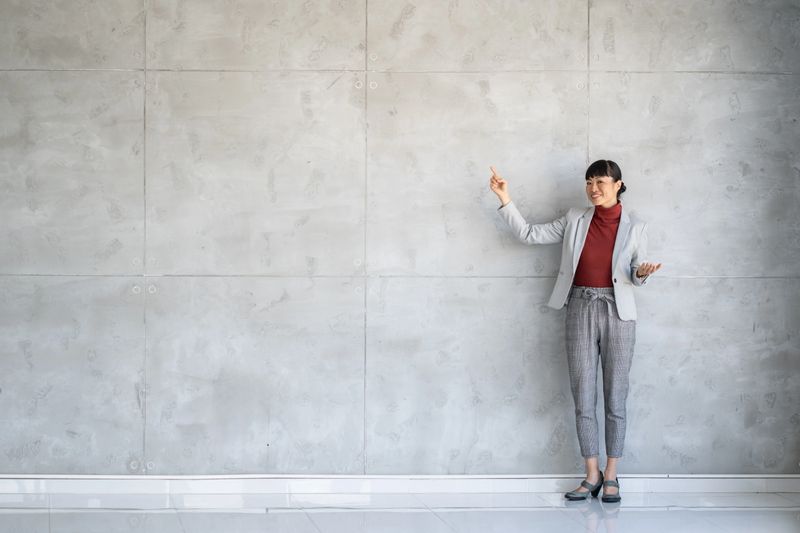 A confident businesswoman stands against a modern concrete wall, gesturing as if presenting an idea or concept, showcasing professionalism.