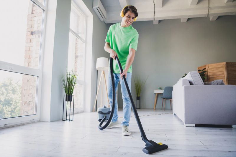 A young man happily vacuuming the stylish and bright interior of a living room, dressed casually and enjoying his cleaning routine in a comfortable and homey atmosphere.
