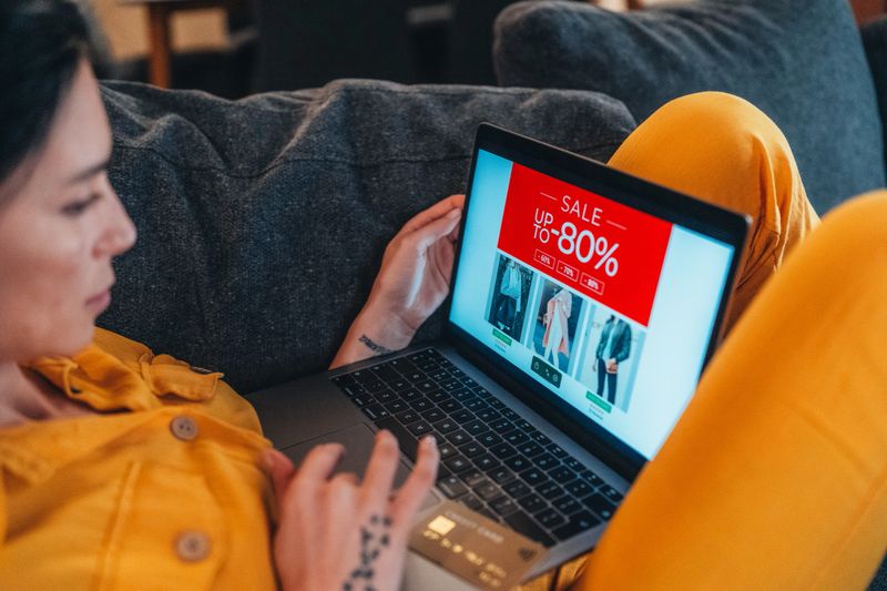 The image features a person holding a credit card while intently looking at a laptop. The laptop screen displays the word "SALE" with discounts ranging from 60% to 80%. Dressed in bright yellow clothing, the individual exudes enthusiasm and excitement about online shopping. This scene captures the essence of modern consumerism and the thrill of finding great deals, making it ideal for themes related to e-commerce, savings, and digital shopping experiences.