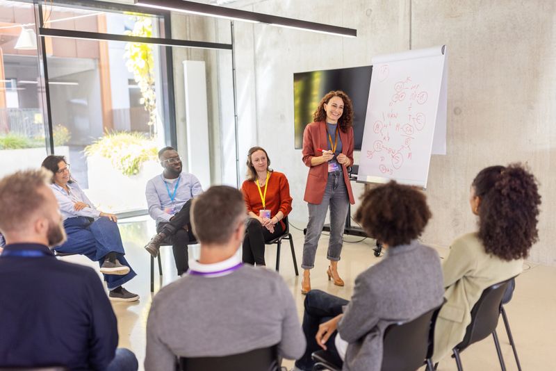 Businesswoman presenting new business ideas on a flipchart to a diverse group of professionals seated in a circle. Showcases her ability to clarify concepts and foster teamwork in a collaborative setting.