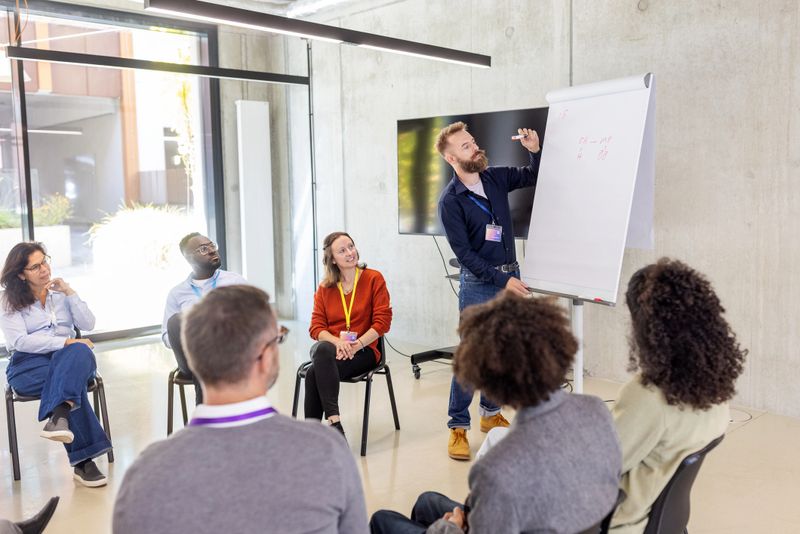 Businessman writing on a flipchart while giving a presentation to multiracial businesspeople seated in a circle. Highlights collaborative discussions and effective communication at a conference hall.