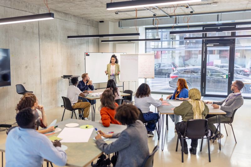 Female professional standing by a flipchart, addressing businesspeople seated in small groups at different tables. Highlights her leadership skills and interactive approach during a seminar at a convention center.