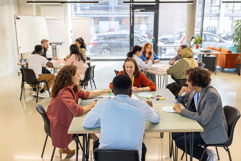 Group of multiracial professionals seated at tables, actively involved in seminar tasks. Reflects teamwork, engaging interactions, and effective collaboration in a professional convention setting.