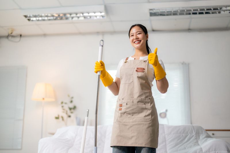 Asian female cleaning professional wearing yellow gloves, holding mop, giving thumbs up with cheerful smile