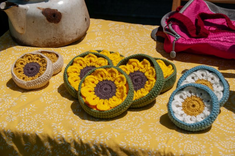A collection of vibrant crocheted sunflower and daisy motifs arranged on a yellow patterned tablecloth, capturing a cozy and creative outdoor setting with handmade artistry pieces on display.