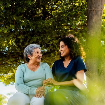 A caregiver warmly supports an elderly woman outdoors under a tree.