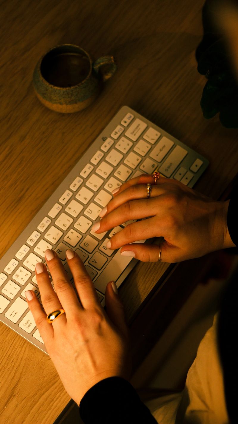 A warm and cozy home office setup featuring a wooden desk with a wireless keyboard and a rustic ceramic coffee cup. The image captures the hands of a woman elegantly typing, adorned with stylish gold and gemstone rings. The soft, ambient evening light enhances the intimate and productive atmosphere, highlighting the balance between work and comfort. Perfect for themes of remote work, freelance lifestyle, and creative professionalism.