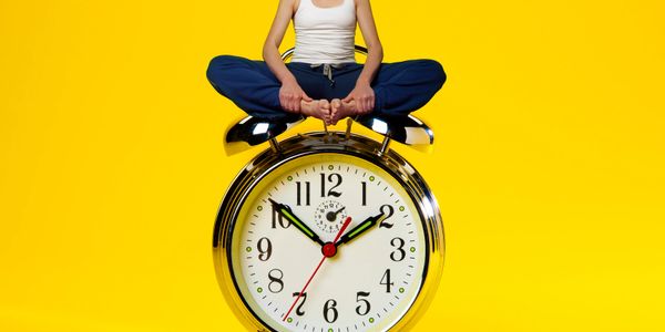 A woman sitting cross-legged on a giant alarm clock against a yellow background.