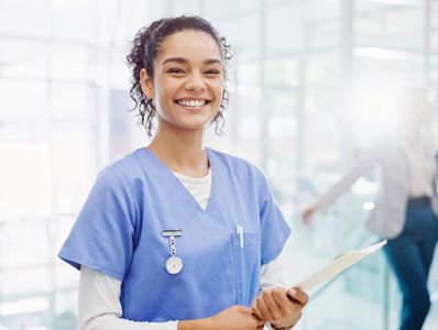 Smiling nurse in blue scrubs holding a clipboard in a bright hospital corridor.