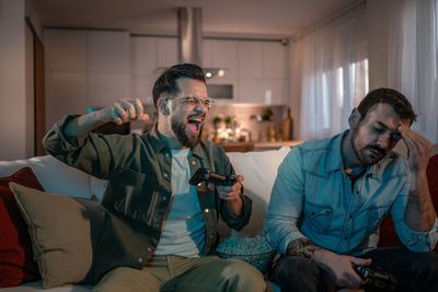 2 men sitting on a couch playing a game. One raises his fist in victory. The other, face-palm.