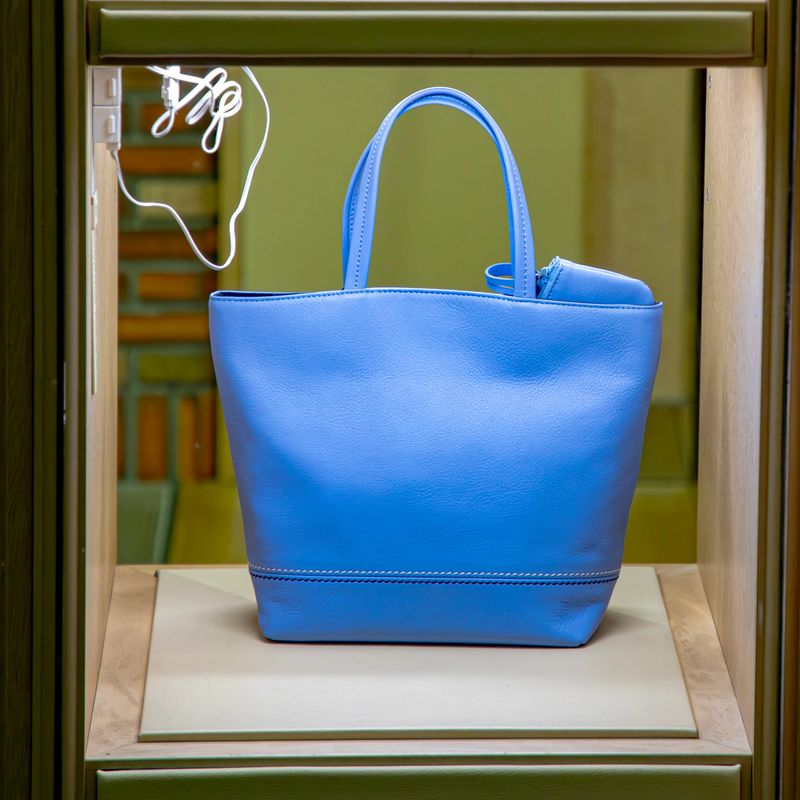 A shot of a blue leather tote bag displayed in a store window. The bag is positioned on a light-colored platform inside a display case with a dark green frame. The bag has two handles and a small pouch attached to one side. The leather appears smooth and the bag is structured. The image captures a retail display of a stylish leather bag.