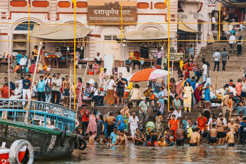 The image portrays Dashashwamedh Ghat by the Ganges River in Varanasi, India, showcasing people gathered for activities including rituals and bathing, embodying the spiritual and cultural richness of this iconic location.