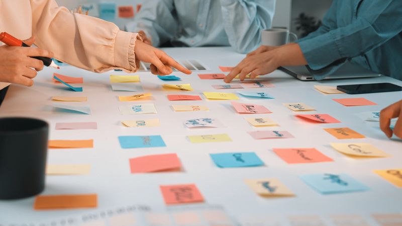 A vibrant scene of diverse hands reaching for colorful sticky notes on a white table, highlighting creative collaboration and brainstorming in a modern workspace setting. SACTR
