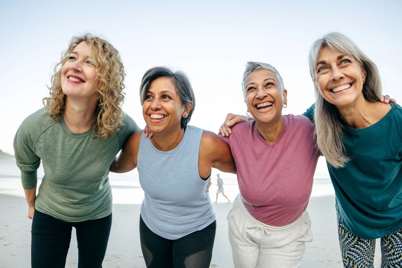 A group of middle-aged sporty women stands together on the beach, dressed in athletic wear. They lean in with arms around each other, exuding energy, friendship, and an active lifestyle. Their bright expressions reflect joy and camaraderie against the serene coastal backdrop.