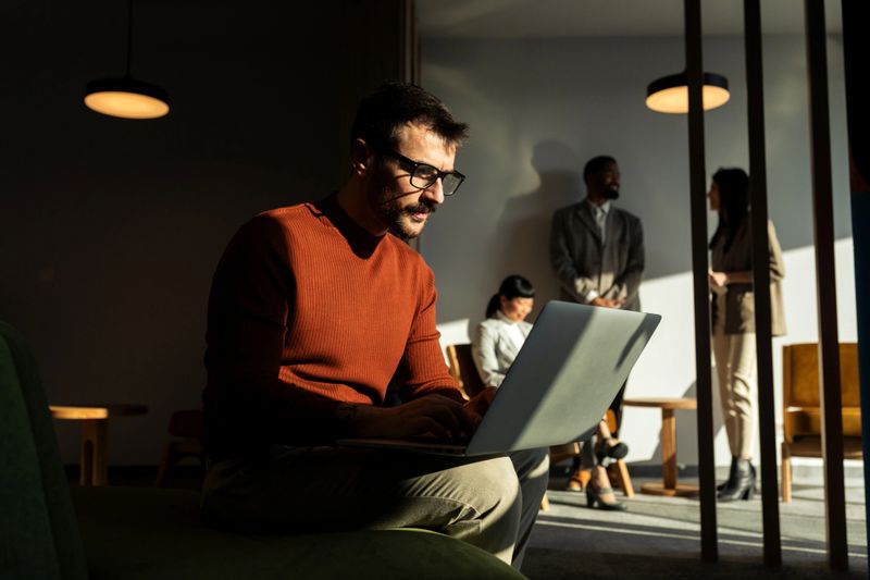 Mid adult man, sitting in a open space office, working on laptop computer, having online meeting while his colleagues have brainstorming in the background