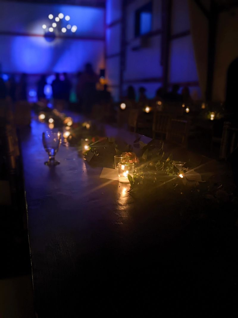 Rustic barn wedding or event reception featuring warm lighting, elegant wooden tables, candles, decor accents, and subtle blues with people dancing in the background. Intimate atmosphere highlights charm and welcoming ambiance.
