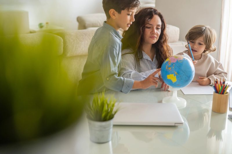 Mother assisting her two sons with their geography homework, pointing at locations on a globe while they sit at a table with a laptop and colored pencils