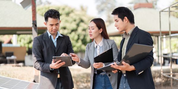 Three professionals discuss work outside with tablets and documents.