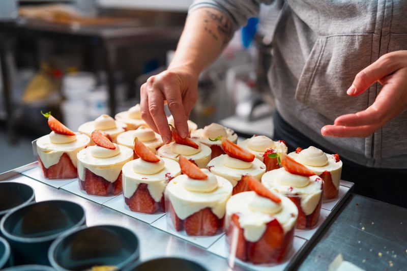 Professional pastry chef garnishing mini strawberry cakes, carefully decorating delicate desserts with precision and skill in commercial kitchen setting