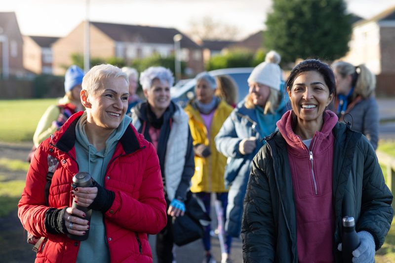 A waist-up shot of a group of mature women dressed in warm, sporty attire arriving to netball training together. They are smiling and carrying water bottles. Located in a suburban park area, with houses and greenery in the background, illuminated by bright morning sunlight. 

Videos of this scenario are available