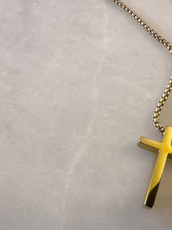 Close-up of a gold cross pendant on a chain against a light background.