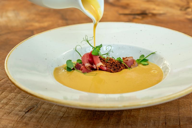 A fine dining dish featuring a creamy gourmet soup being poured into a deep white plate. The dish is elegantly garnished with smoked meat slices, microgreens, and crispy crumbles, creating a visually appealing and luxurious presentation.
