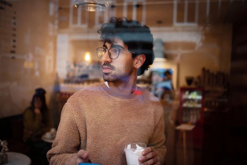 A casual man with glasses enjoying a drink in a cozy cafe, seen through a reflective window, showcasing a thoughtful and serene atmosphere.
