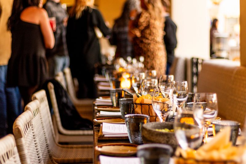 A long dining table elegantly set with glassware, plates, menus, and appetizers, ready for a lively gathering as guests mingle in the background.