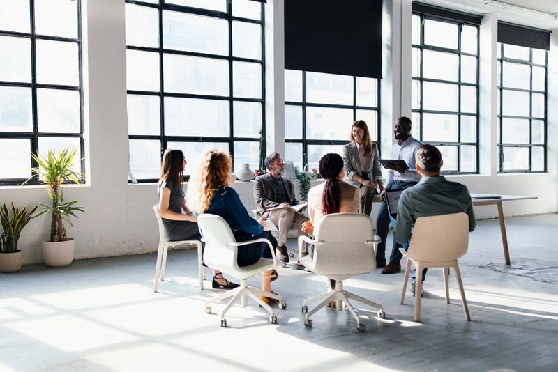 Diverse professionals collaborate in a meeting within a modern bright office. They sit together in a circle, promoting teamwork, creativity, and effective communication in a co-working business environment.