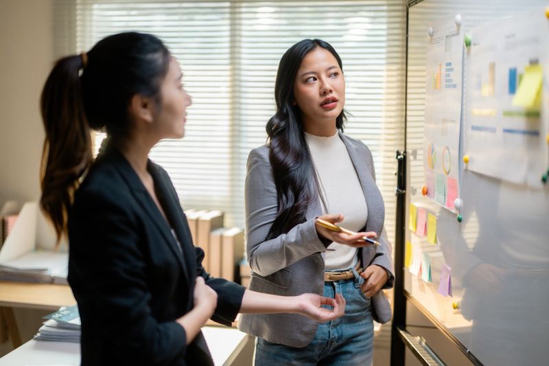 Professional women collaborating, reviewing business analytics near whiteboard with graphs, discussing strategic planning during workplace meeting