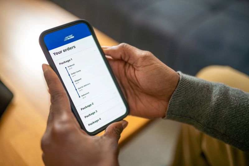 A close-up view of hands holding a smartphone showing a shipping order tracking screen. The scene is indoors, well-lit, and features a casual background with a wooden table and soft furnishings.
