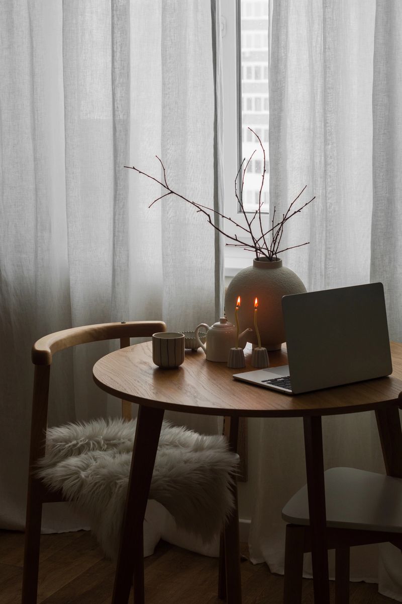 The cozy evening interior of the living room - a round wooden table with a ceramic vase, a laptop, a cup, burning candles and minimalist chairs by the window