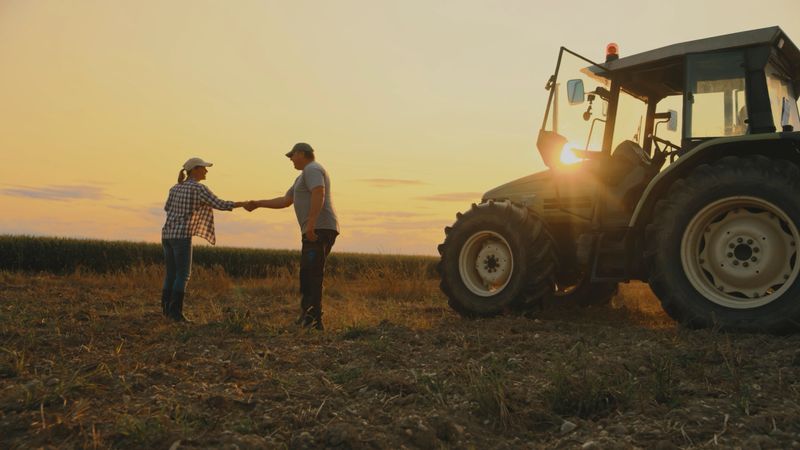 Man and a Woman,Both Farmers,Shake Hands in a Field Next to a Large Tractor at Sunset. The Gesture Suggests Agreement,Partnership,Or the Completion of a Successful Collaboration
