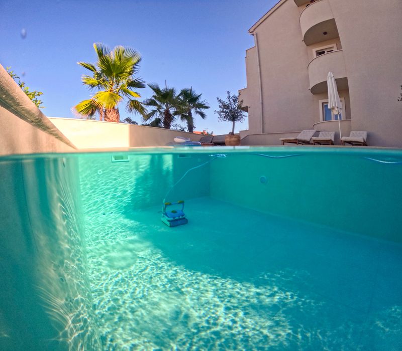 A serene outdoor villa pool being cleaned by a robotic cleaner, surrounded by palm trees and modern architecture under a sunny blue sky.