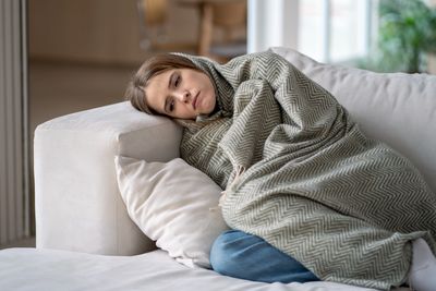 A woman wrapped in a blanket resting on a couch looking tired.