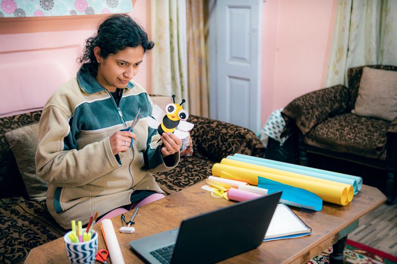 A female primary school teacher in a beige fleece jacket works on an arts and crafts project at home. She learns craft skills online using her laptop to teach her students. Her home workspace features a wooden table with colorful craft materials, a mug filled with pens, and a decorated couch in the background.
