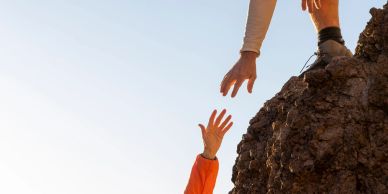 One person reaching to help another climbing a rock.