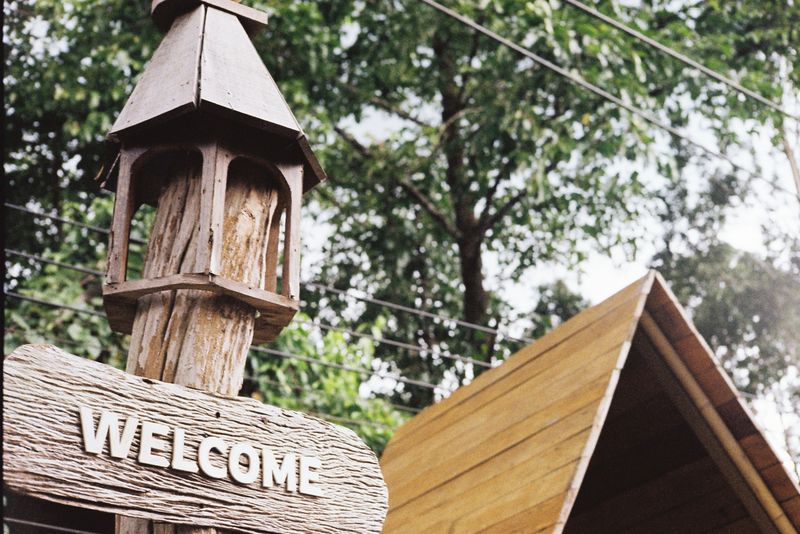 This image gives a sense of warm and friendly welcome. The “WELCOME” sign made of wood with a natural texture reflects the simplicity and sincerity of the place. The old wooden pillars and the wooden structure behind them give a sense of a natural accommodation atmosphere, such as a resort, a cottage, or an eco-tourism attraction.

The backdrop of lush green trees enhances the sense of calm and relaxation, similar to being invited to stay in a warm and friendly place. This image conveys a sincere welcome to guests and gives everyone an opportunity to experience nature and peace.